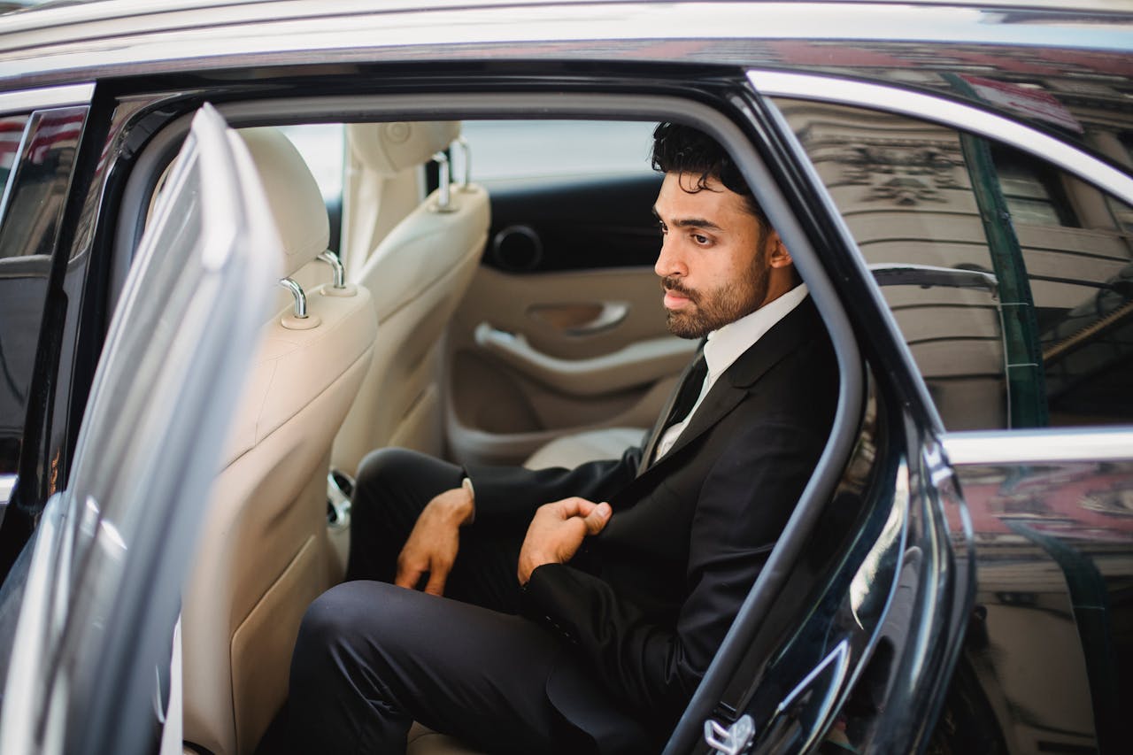 A bearded man in a black suit sits elegantly inside a luxury car, exuding sophistication.